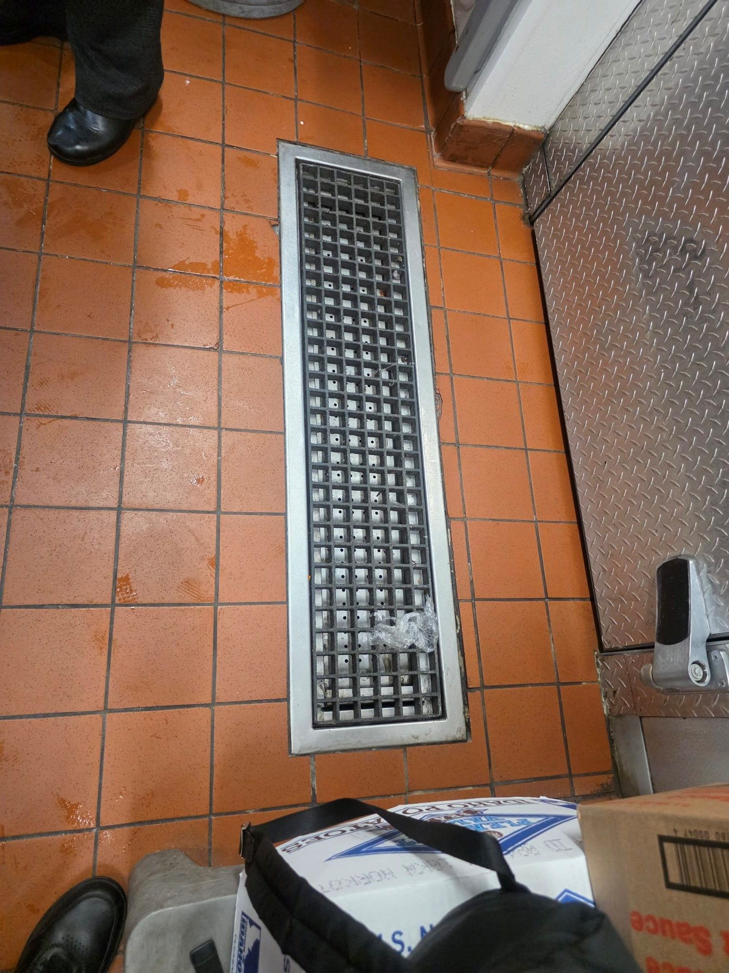Floor vent in a kitchen.  Orange tile floor with a rectangular metal grate.  A person's feet in black shoes are at the edge.