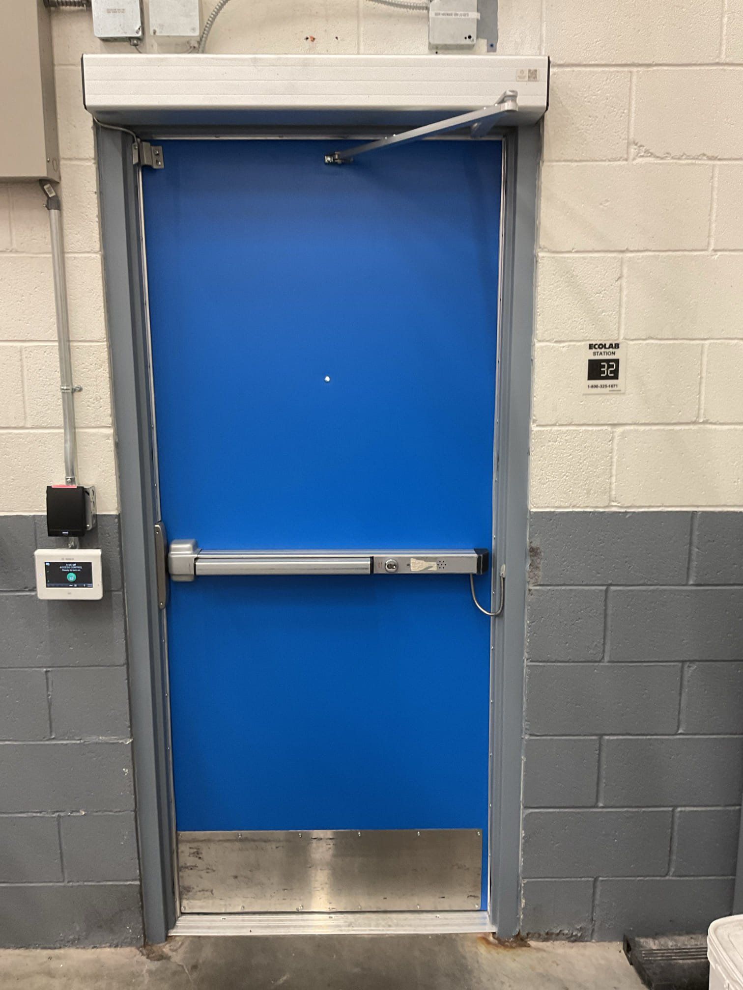 Blue door with a silver bar and hardware, in a gray-brick wall. An automatic closer is at the top.