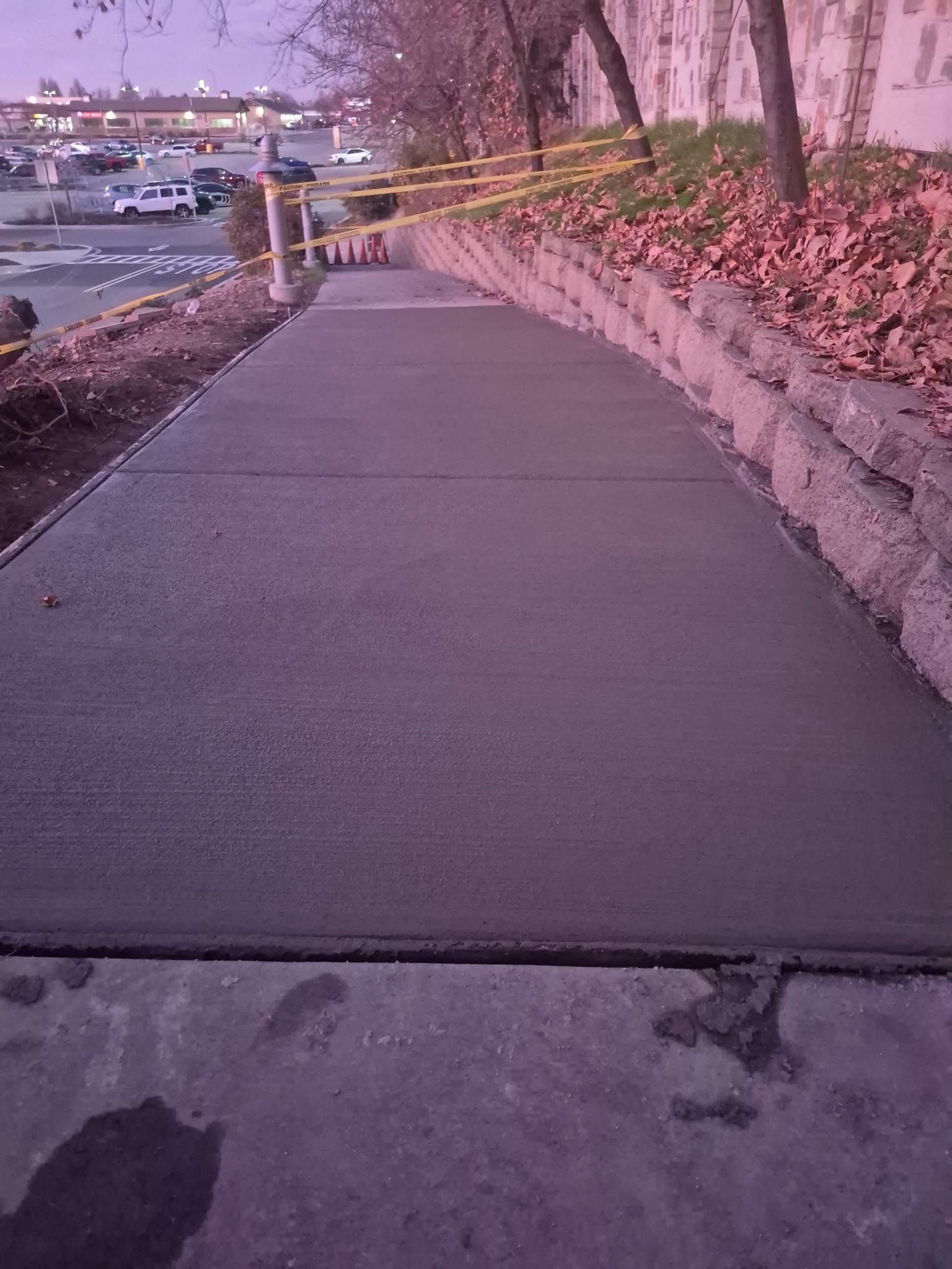 Newly poured concrete sidewalk with caution tape on a sloped path, next to a retaining wall.