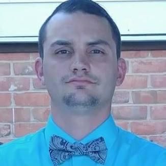 Man in a turquoise shirt and paisley bow tie, in front of a brick wall.