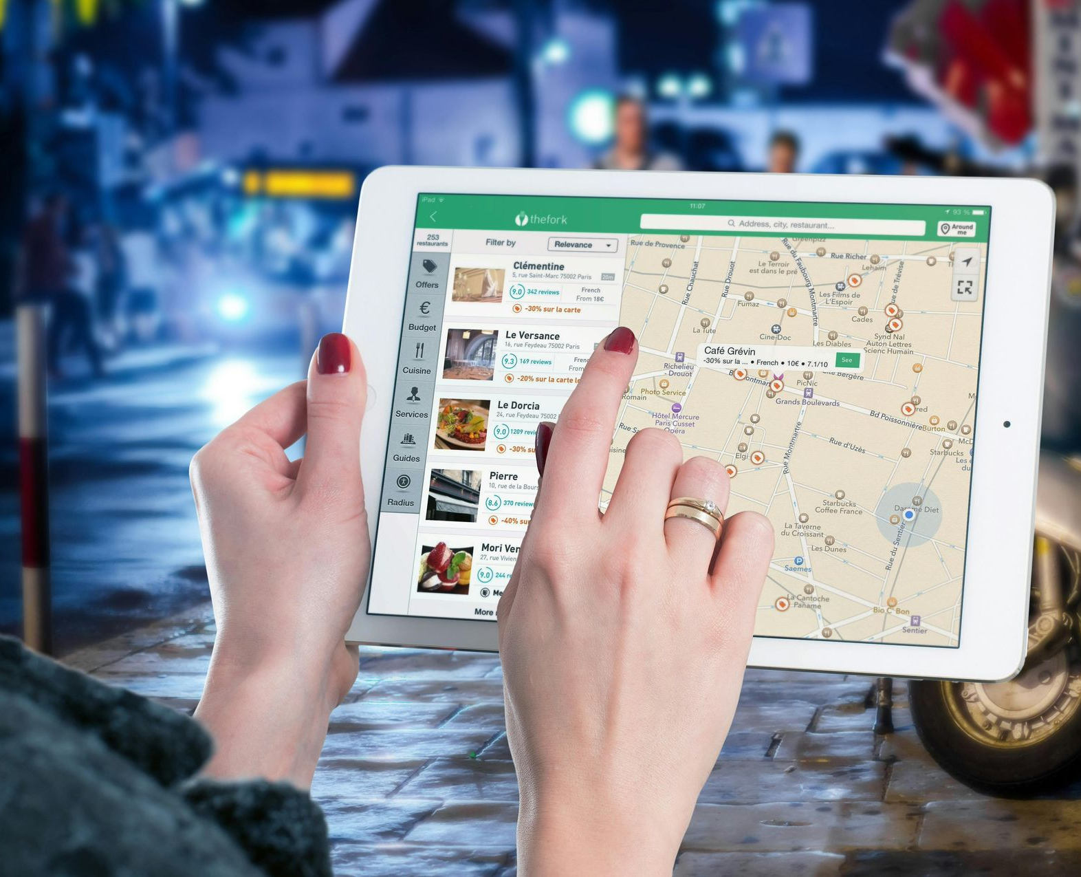 Person holding tablet with map app, using finger to select location on city street at night.