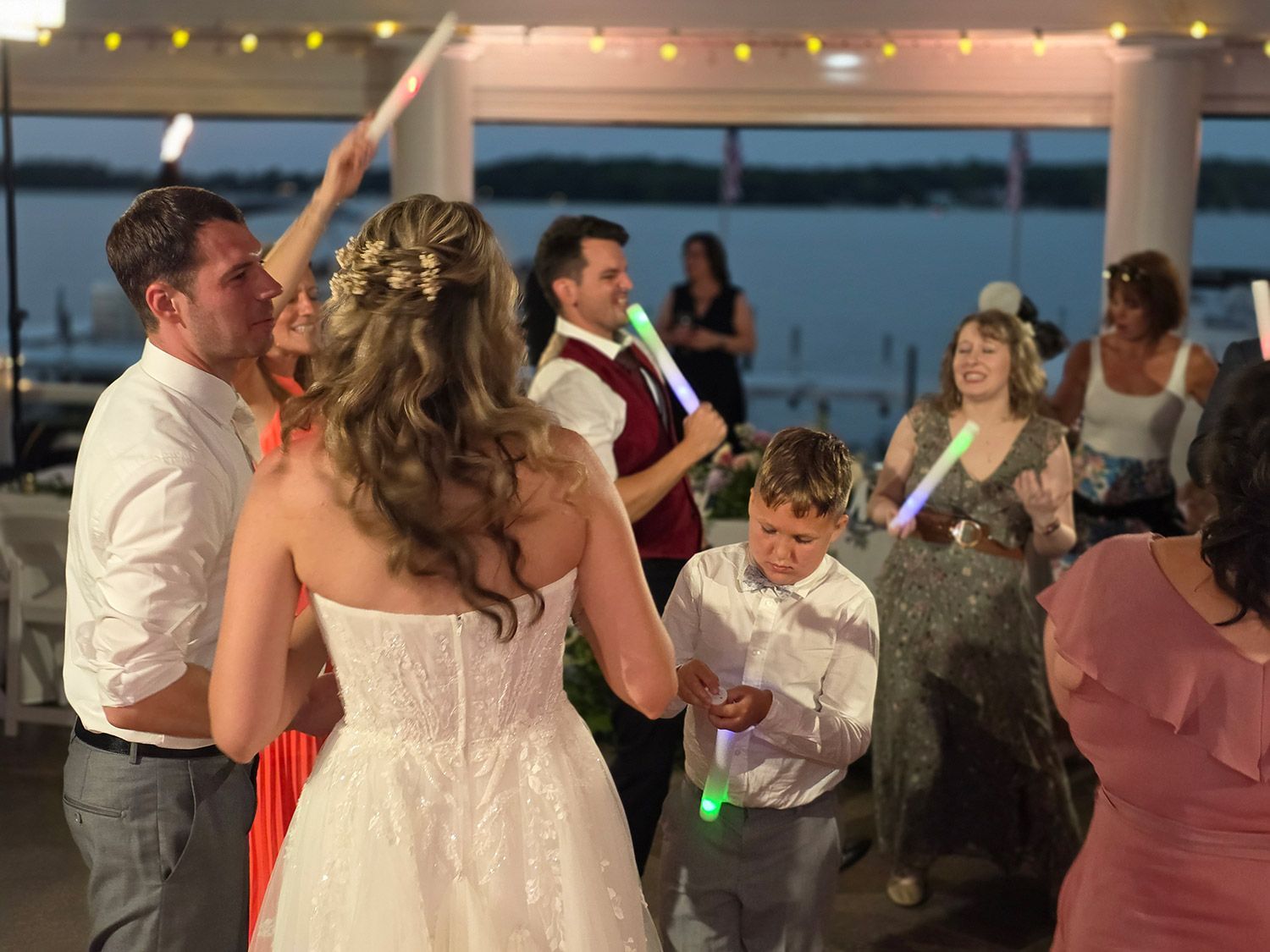 Wedding reception: Bride and groom dance, guests with glow sticks, lake in background.