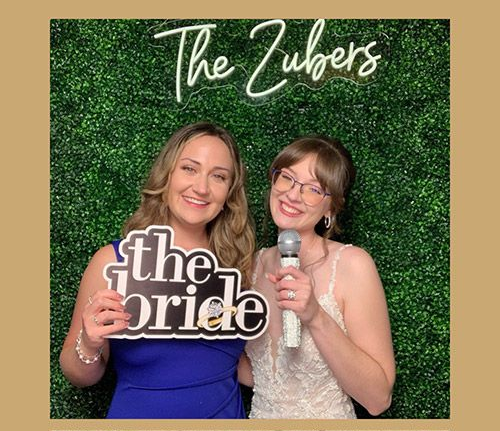 Two smiling women pose at a wedding photo booth. One holds a "the bride" sign; the other a microphone. Green wall, "The Zubers" neon sign.