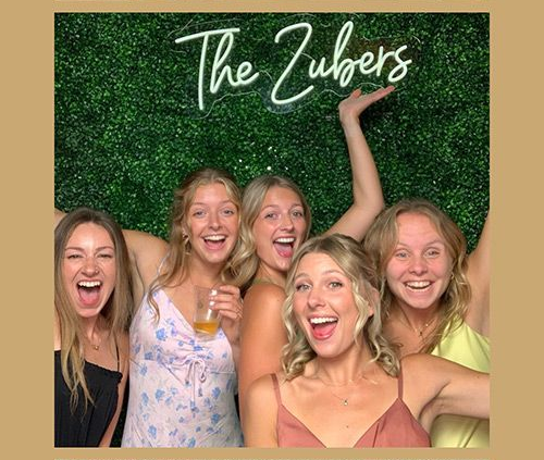 Five excited women pose for a photo booth with "The Zubers" neon sign against a green wall.