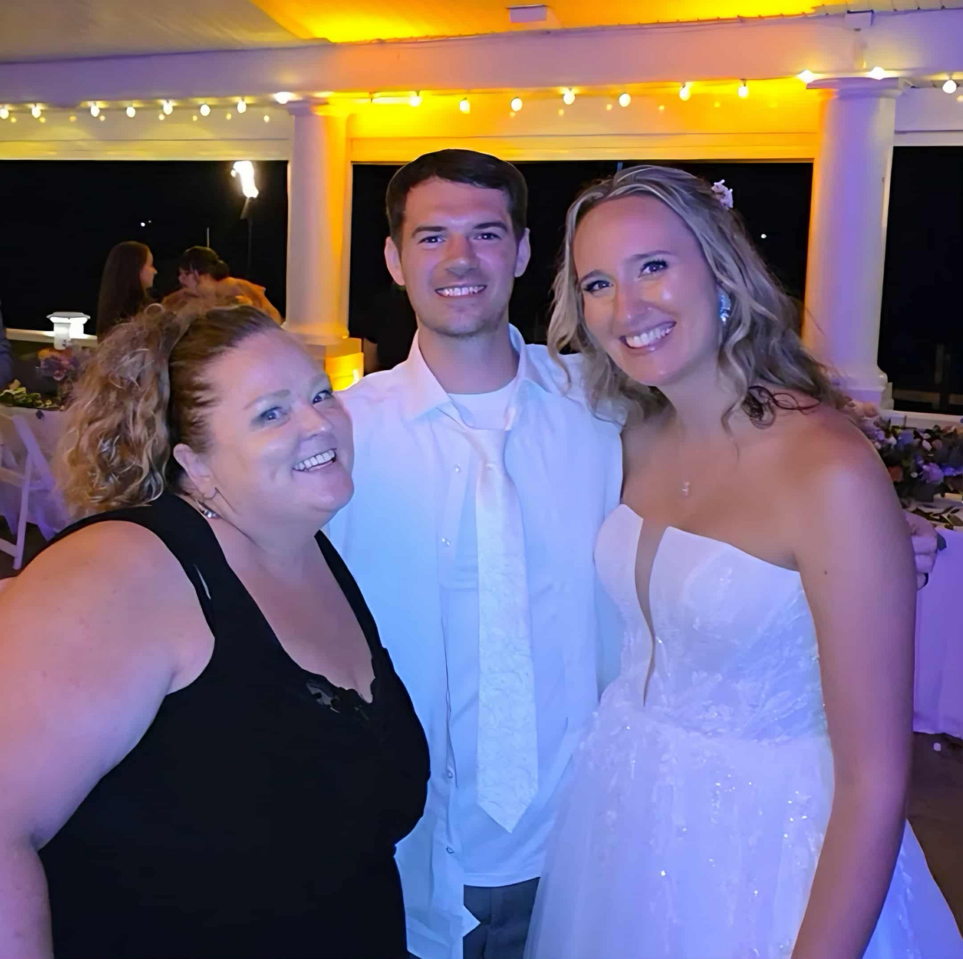 Three people smiling at a wedding, in a pavilion. A bride in a white dress, a man, and a woman.