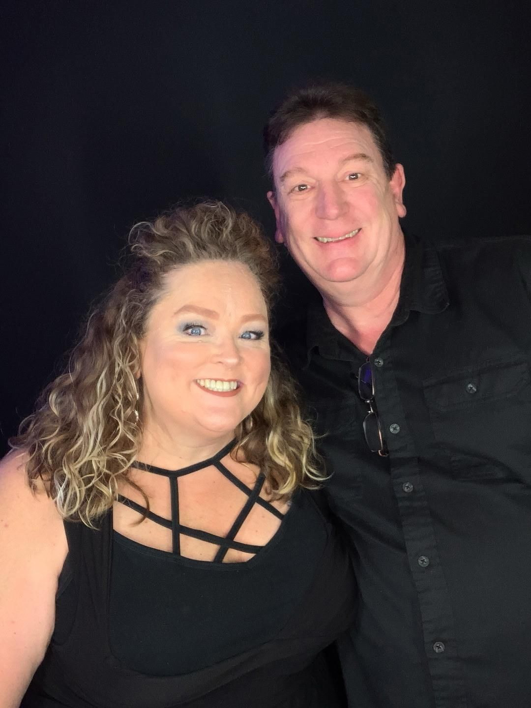 Smiling couple posing for a photo. Woman has curly hair, black top. Man in black shirt. Dark background.