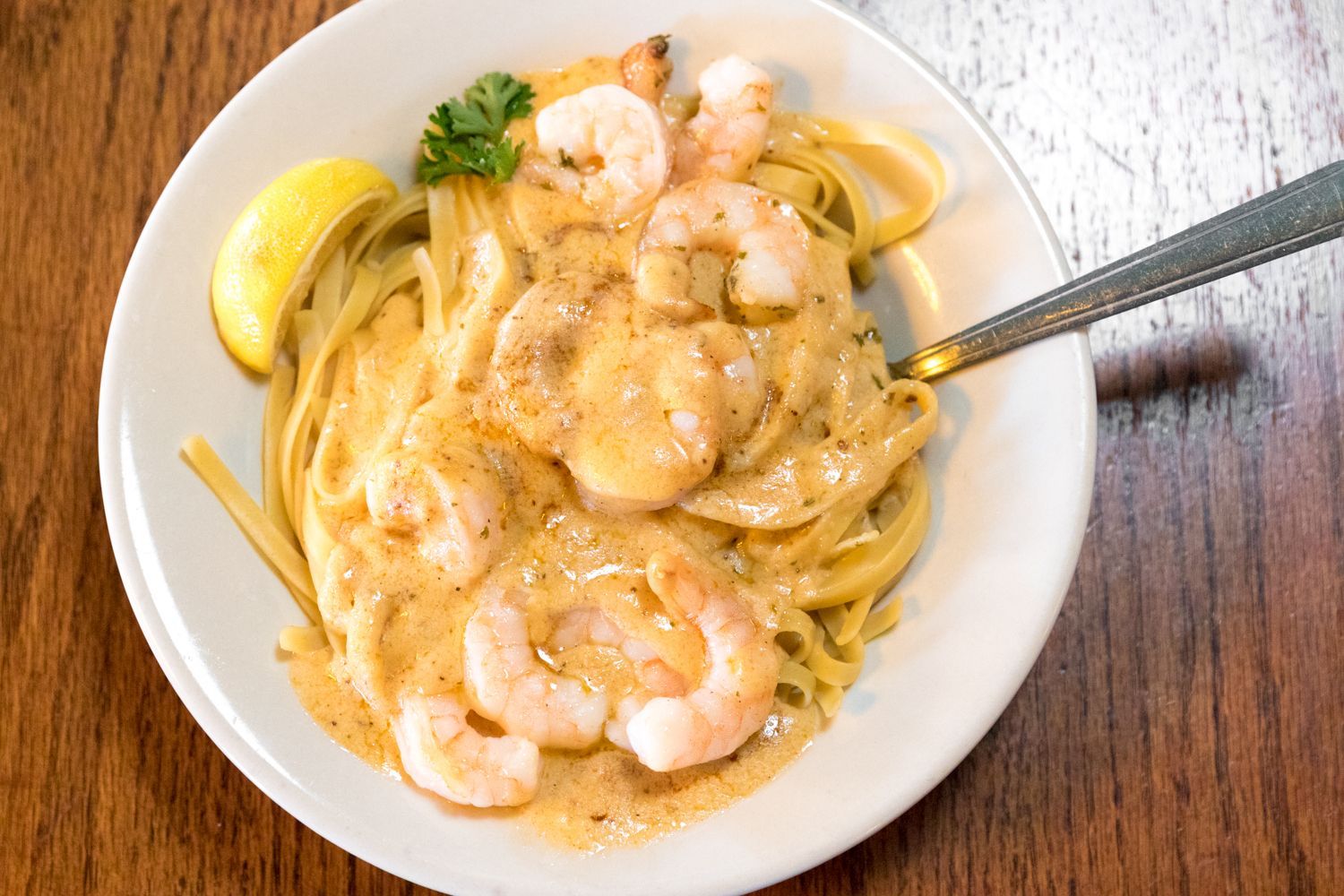 Shrimp scampi with pasta in a white bowl, garnished with lemon and parsley on a wooden table.