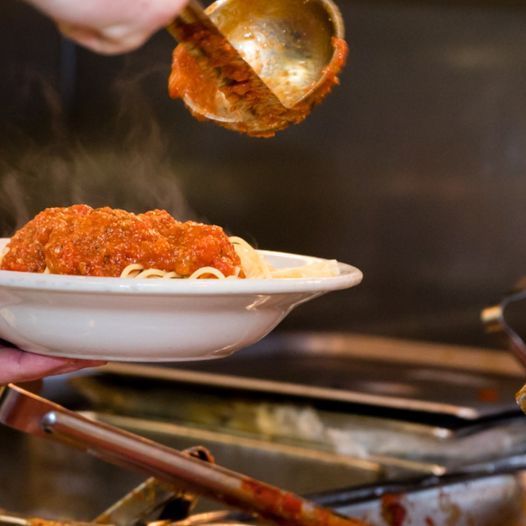 Person ladles tomato sauce onto spaghetti in a white bowl, setting it on a stainless steel counter.