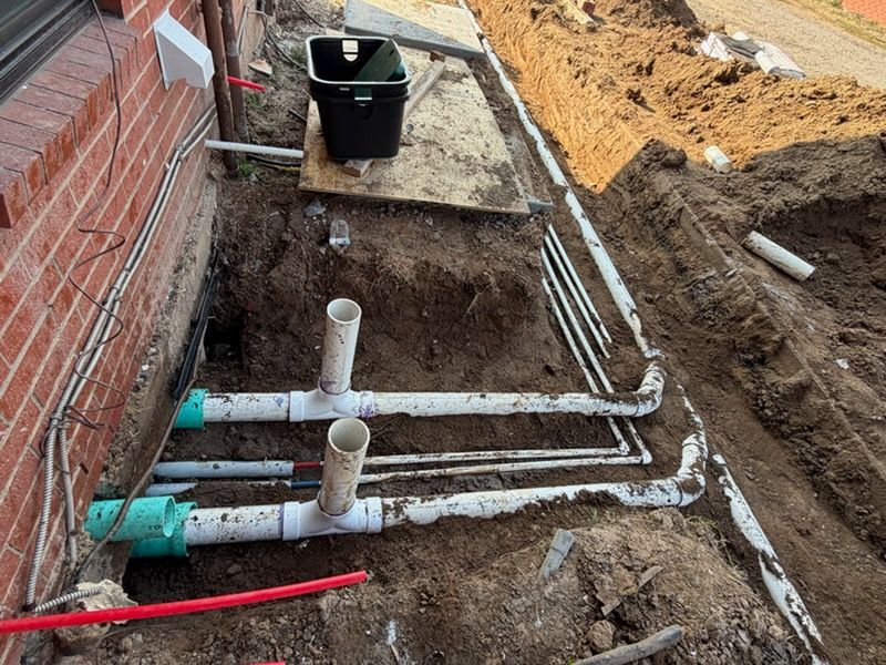 Trench beside brick wall with white PVC plumbing pipes and a black utility box at a construction site