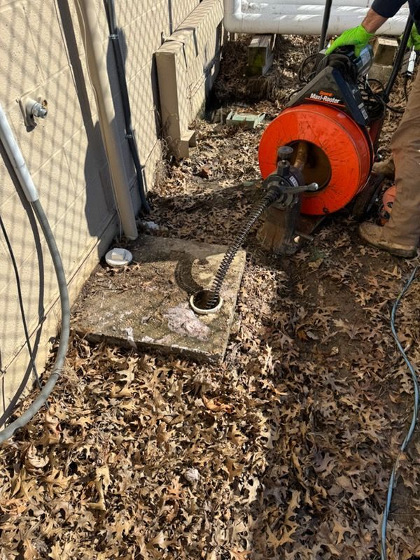 Worker using a drain snake on a clogged outdoor pipe beside a house, with leaves covering the ground.