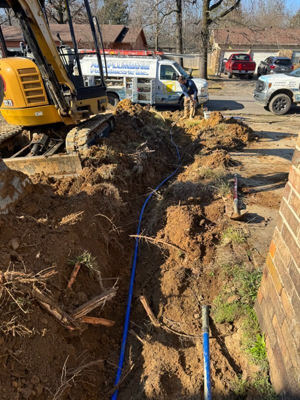 Excavator digging a trench beside a brick wall, with blue utility lines and workers in a muddy yard.