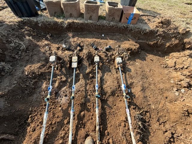 Trenched ground with four irrigation pipes and valves laid in dirt near a row of bricks