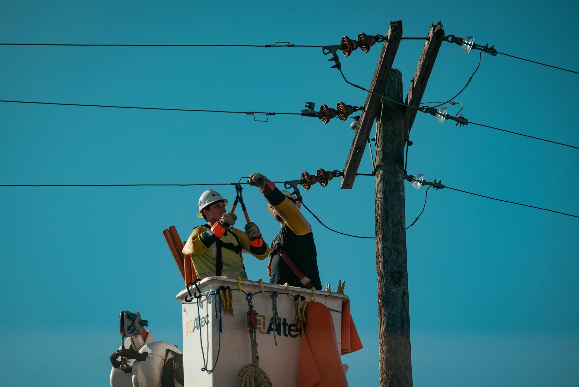 Two utility workers in a bucket truck repair electrical lines on a utility pole against a clear blue sky.