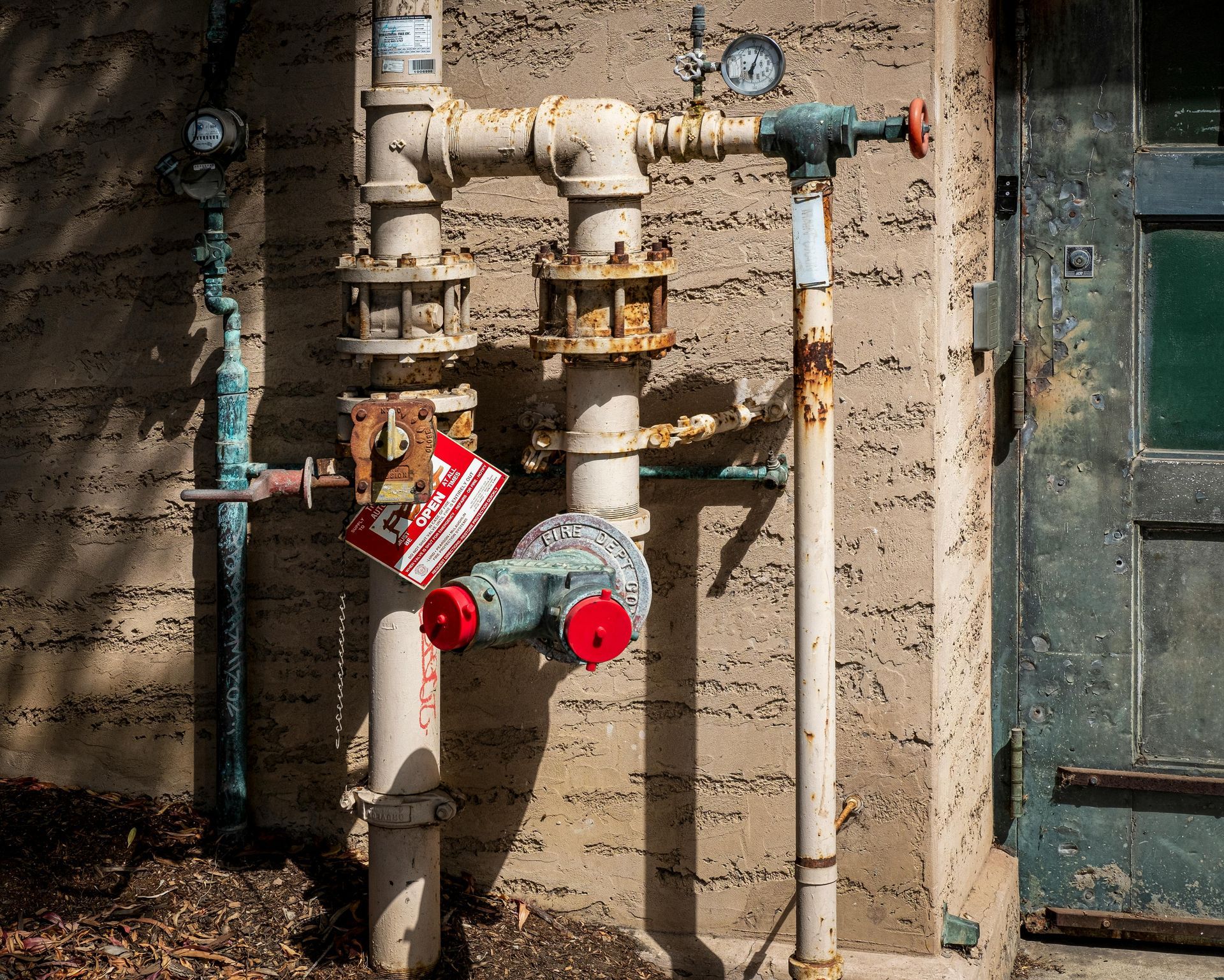 A fire department connection (FDC) with red caps mounted on a tan, textured wall next to a weathered green door.