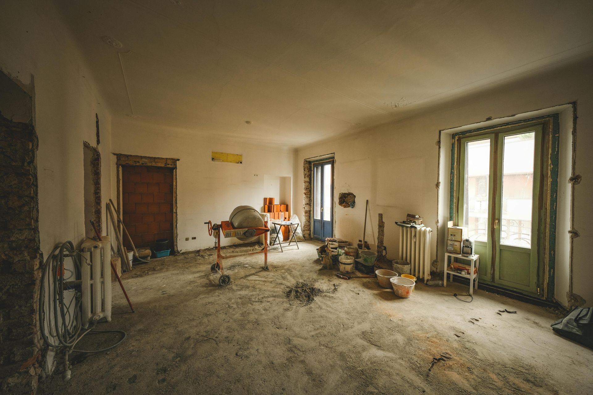A dimly lit, unfinished room undergoing renovation, featuring a concrete mixer, scattered tools, and a pair of glass doors.