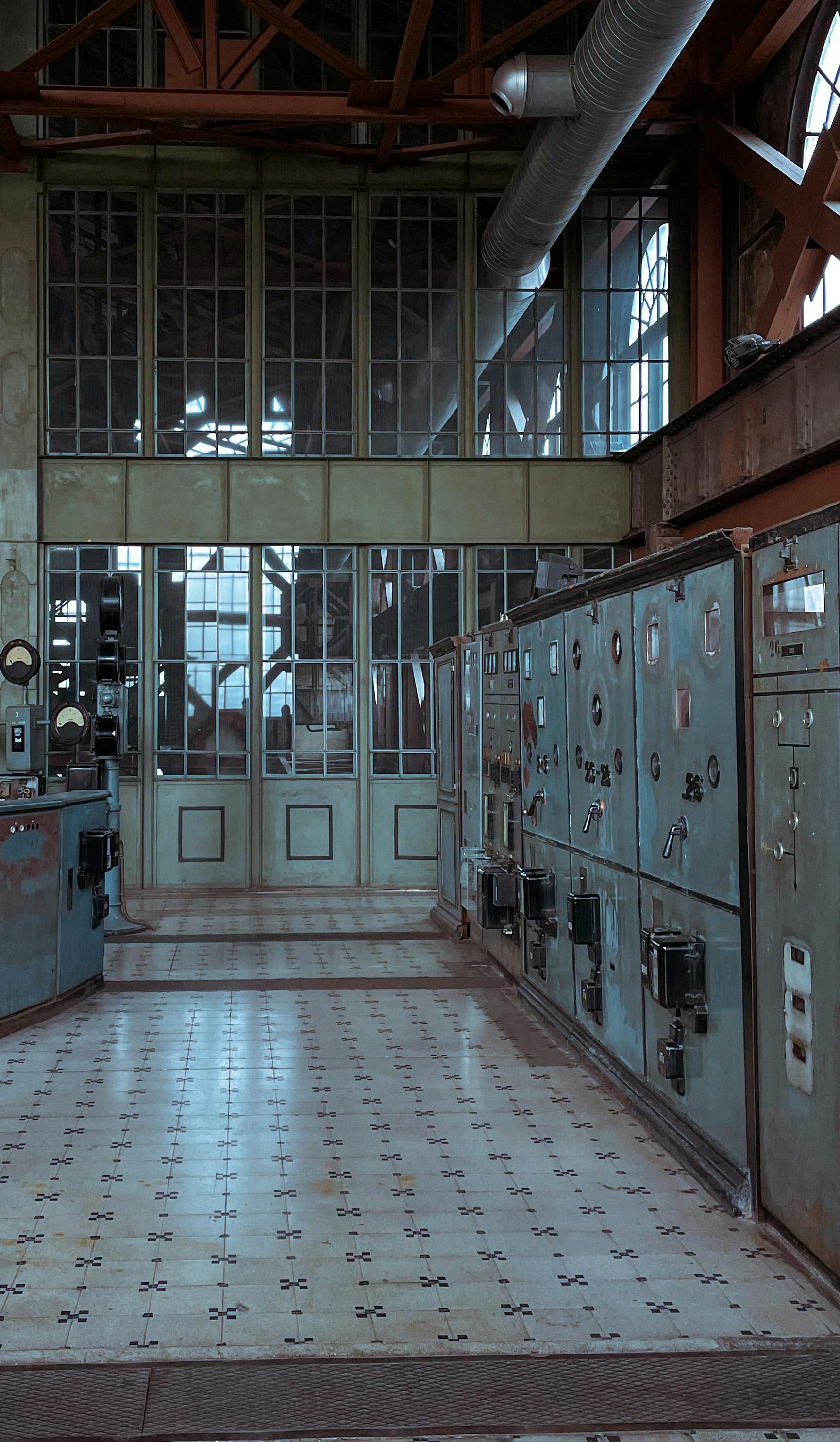 Industrial control room with a grid-patterned floor, metal cabinets, and large windows beneath exposed ceiling beams.