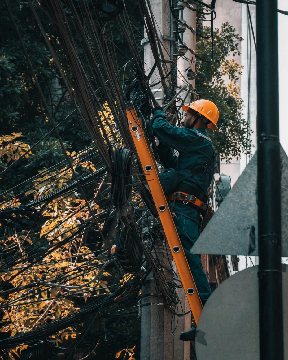 Utility worker in a hard hat and protective gear working on a utility pole while standing on an orange extension ladder.