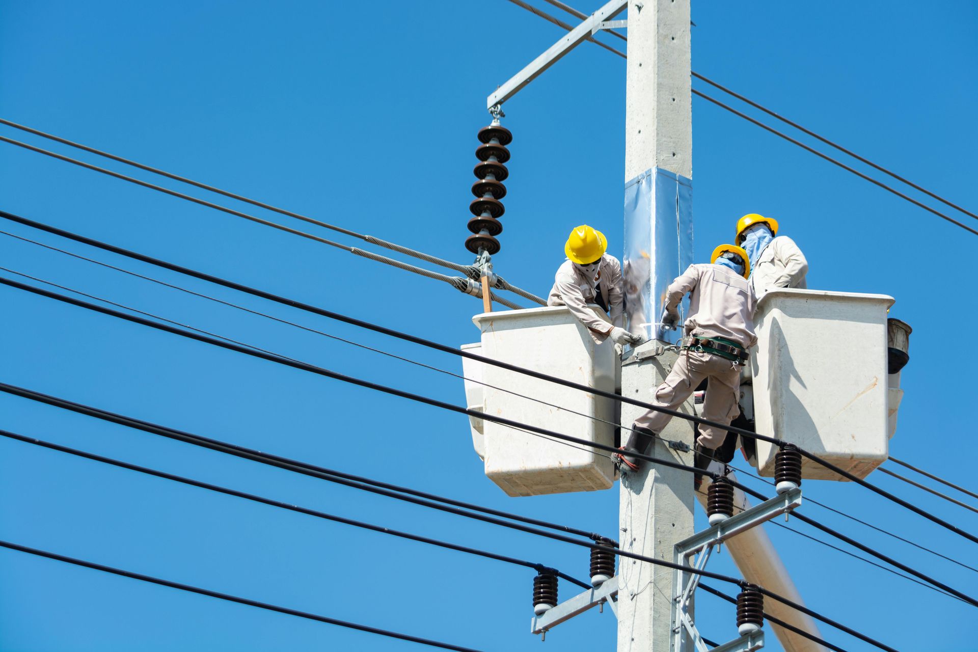 Three utility workers in yellow hard hats and safety gear work from two bucket trucks on a tall power pole.
