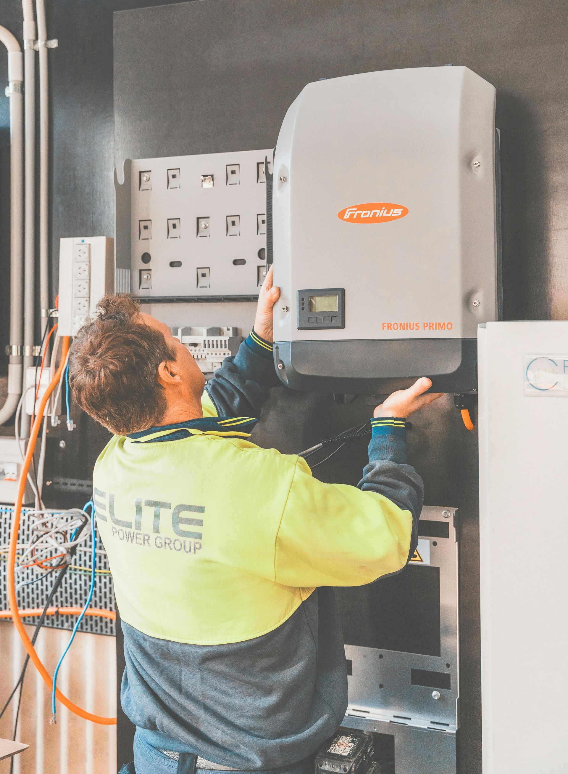 A person in a high-visibility uniform installing a Fronius solar inverter on a wall indoors.