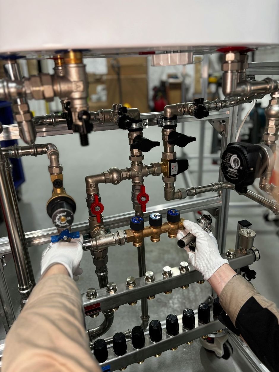 A person in white gloves installs plumbing components and valves on a metal frame of a heating system.