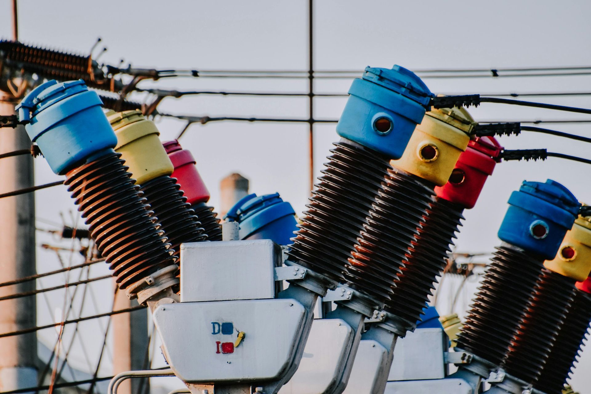Close-up of electrical substation equipment with colorful blue, yellow, and red insulators against a sky background.