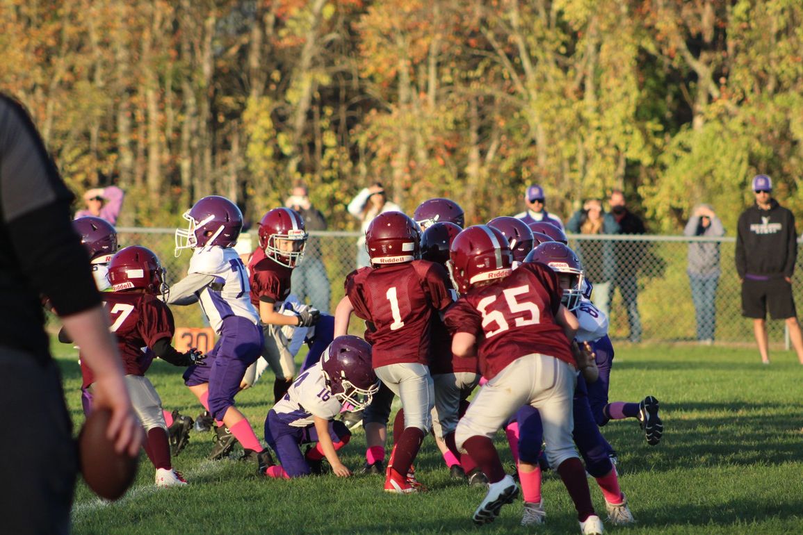 Football players in action on a grassy field; one runs with the ball, pursued by two opponents.