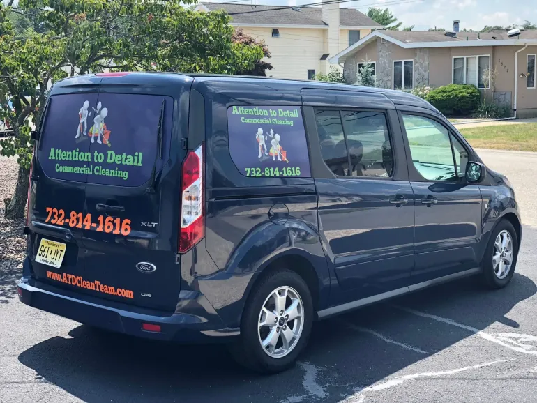 Dark blue Ford van with business logo on windows and phone number. Parked outside buildings.