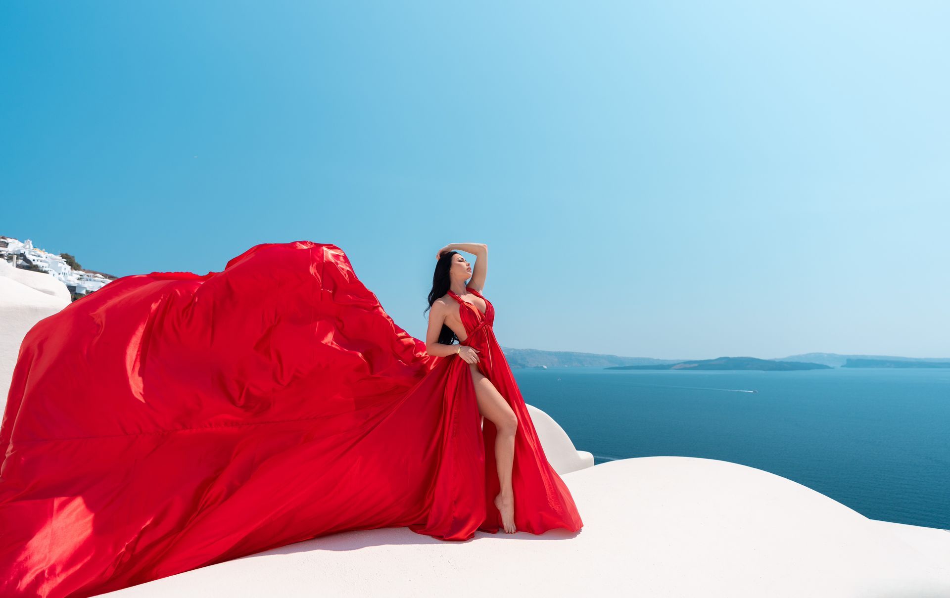 A woman in a long red dress is standing on a snowy hill overlooking the ocean.