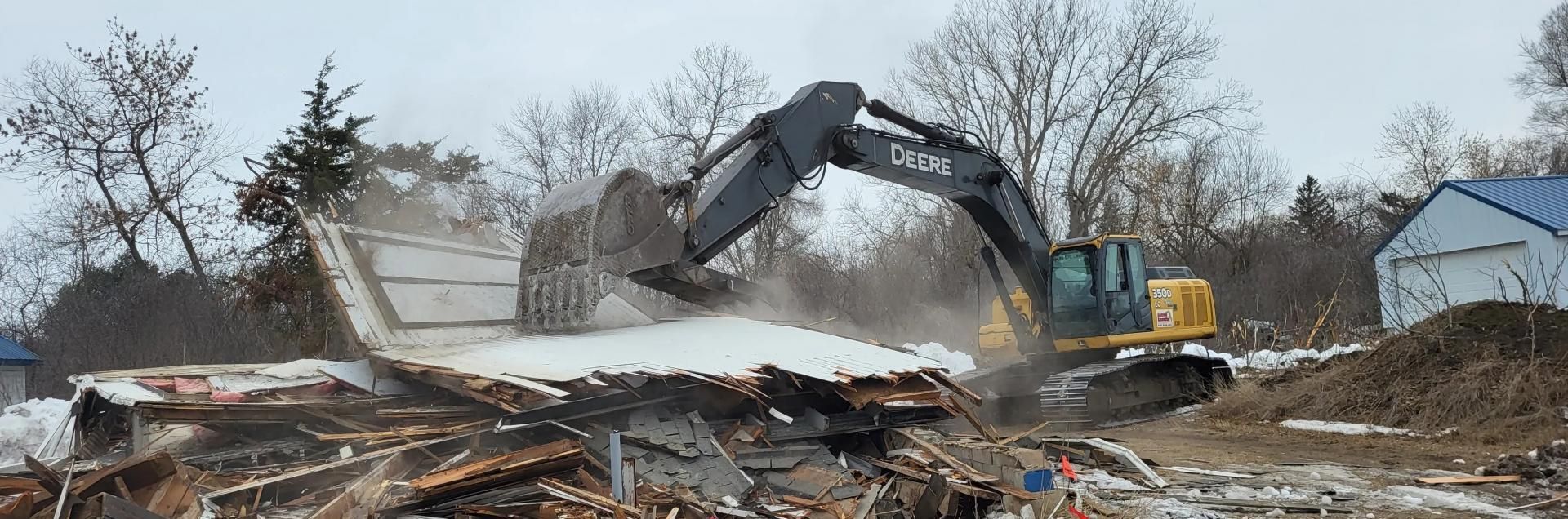 A yellow and black excavator tears down a structure at a demolition site on a cloudy day.