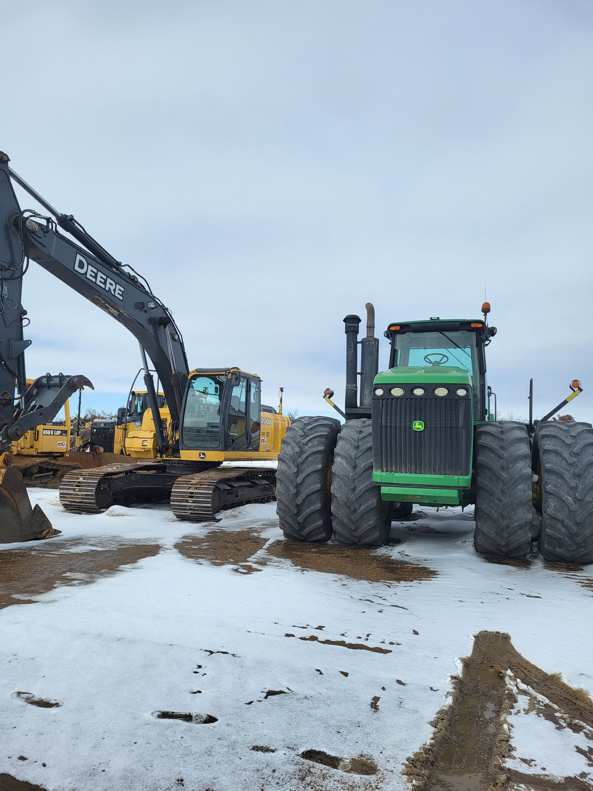 A dark grey excavator with a large arm parked next to a large green tractor with dual tires on snowy, muddy ground.