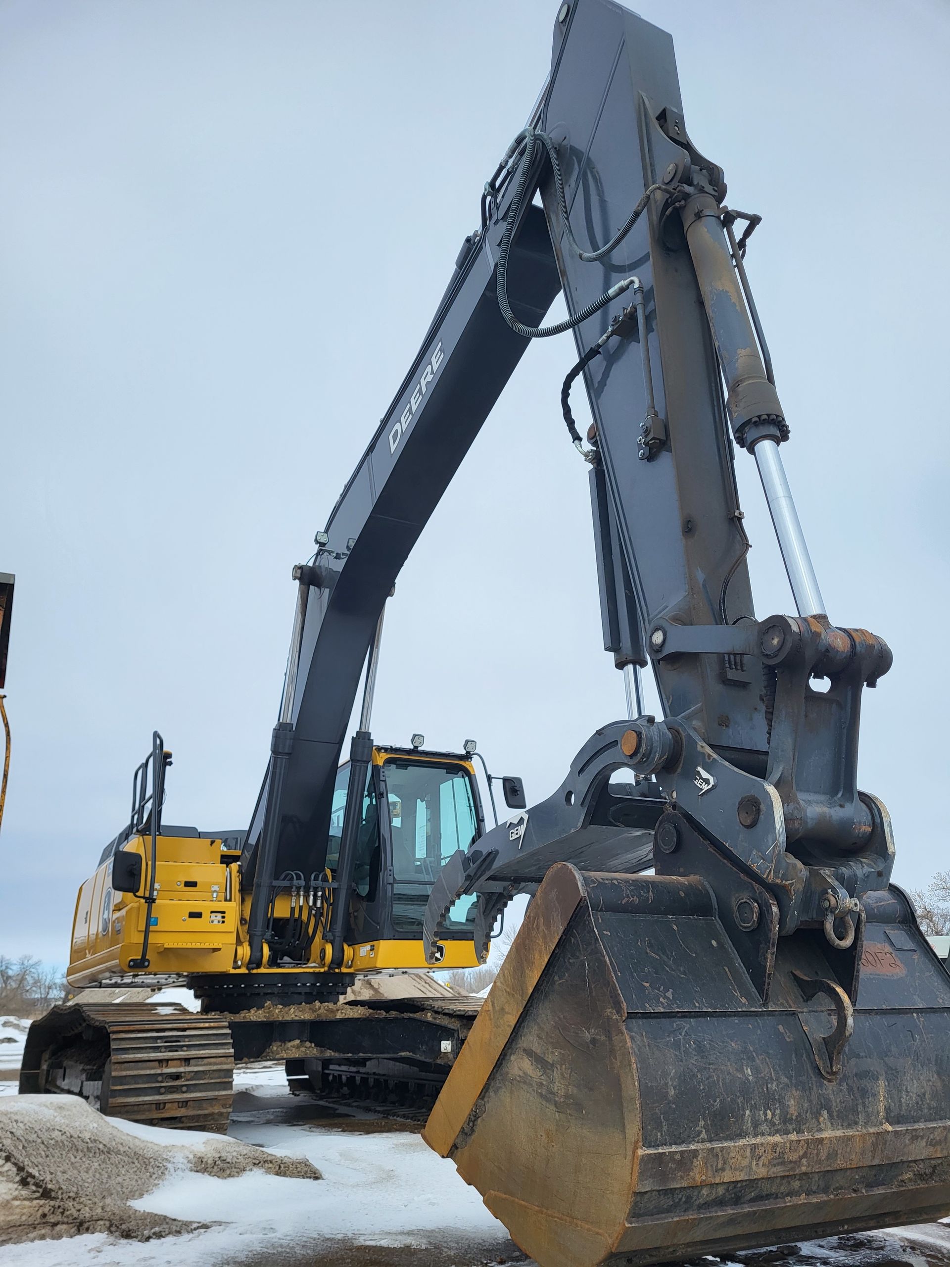 A yellow and grey industrial excavator with a large bucket arm, parked on a snowy ground under an overcast sky.