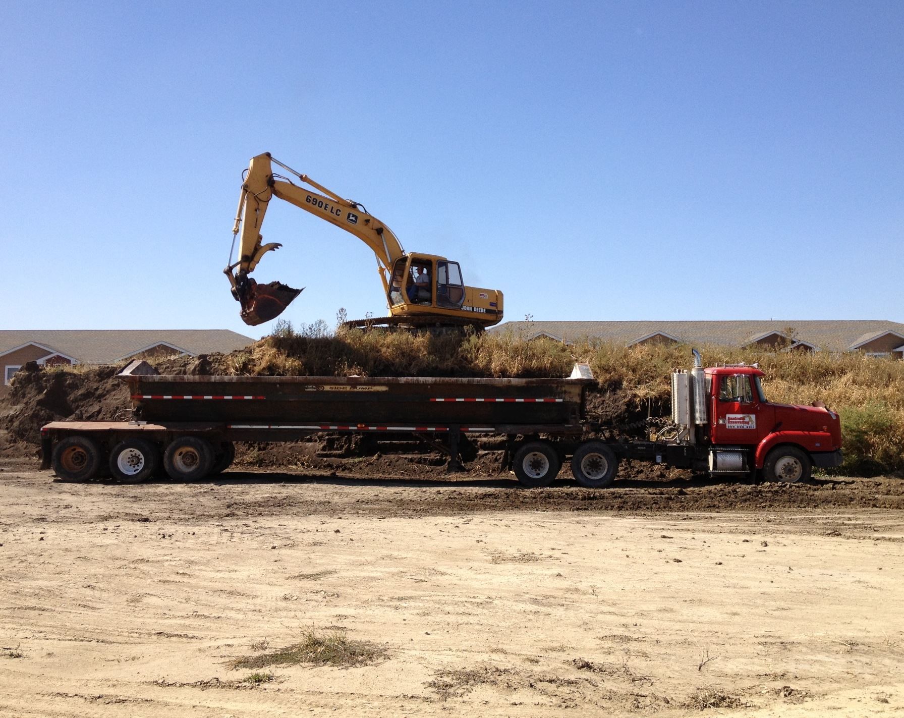 A yellow excavator loads dirt into a red semi-truck trailer on a sunny construction site.