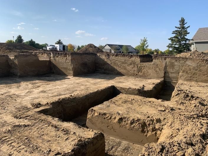 A construction site showing a freshly excavated foundation pit in the ground under a bright, sunny blue sky.