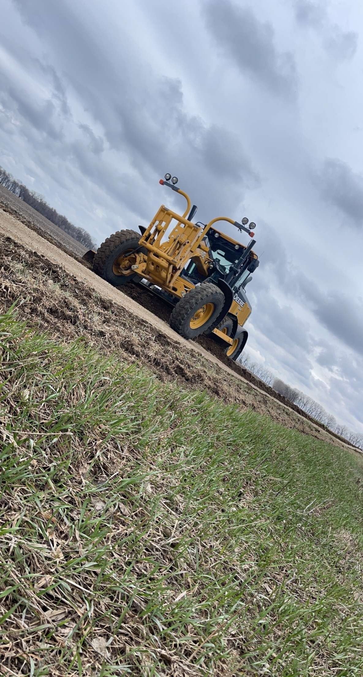 A yellow motor grader works on a dirt path beside a green, grassy field under a cloudy sky.