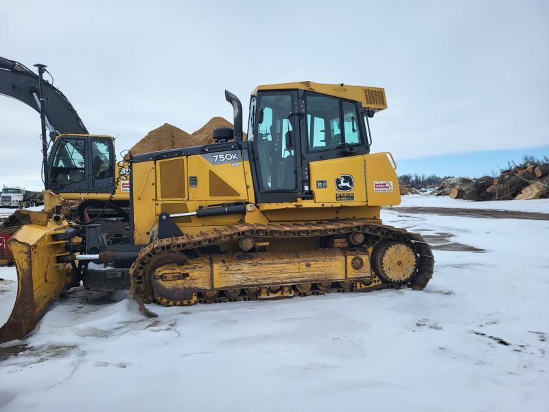 A yellow John Deere crawler dozer sits on a snow-covered ground with an excavator visible behind it.