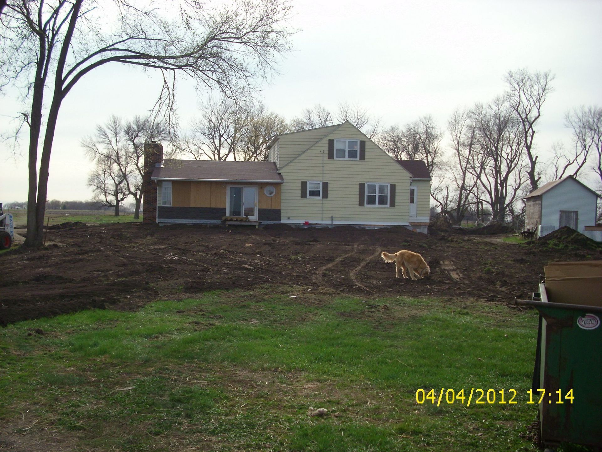 A light-yellow house with a dark roof and chimney sits behind a large, muddy yard with a golden dog walking on the soil.