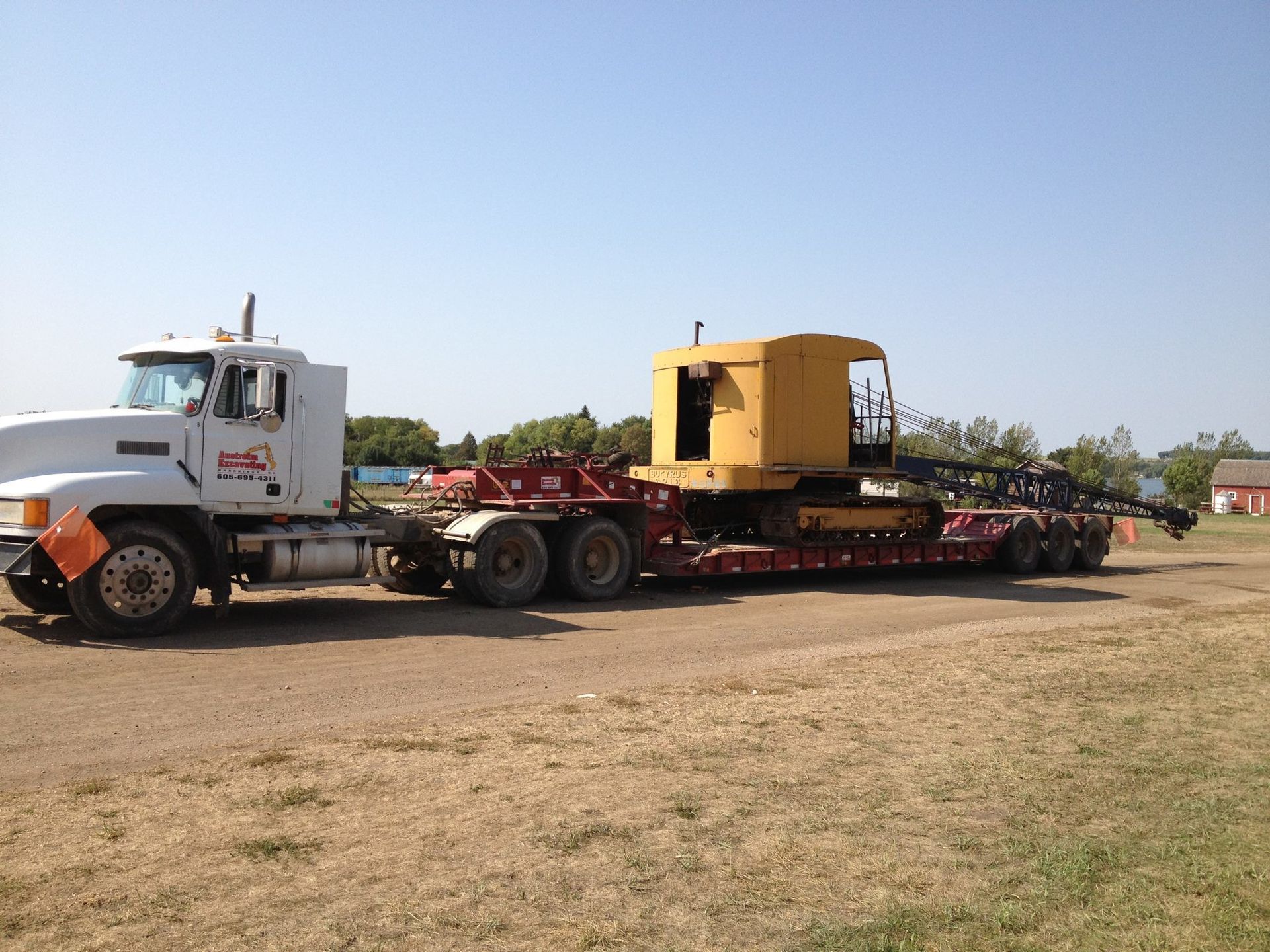 A white semi-truck hauling a yellow heavy construction vehicle on a flatbed trailer across a gravel lot.