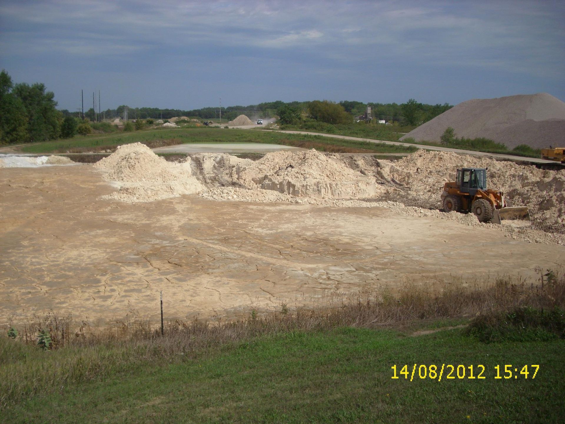 An orange bulldozer parked in a dirt lot next to piles of light-colored crushed rock under a cloudy sky.