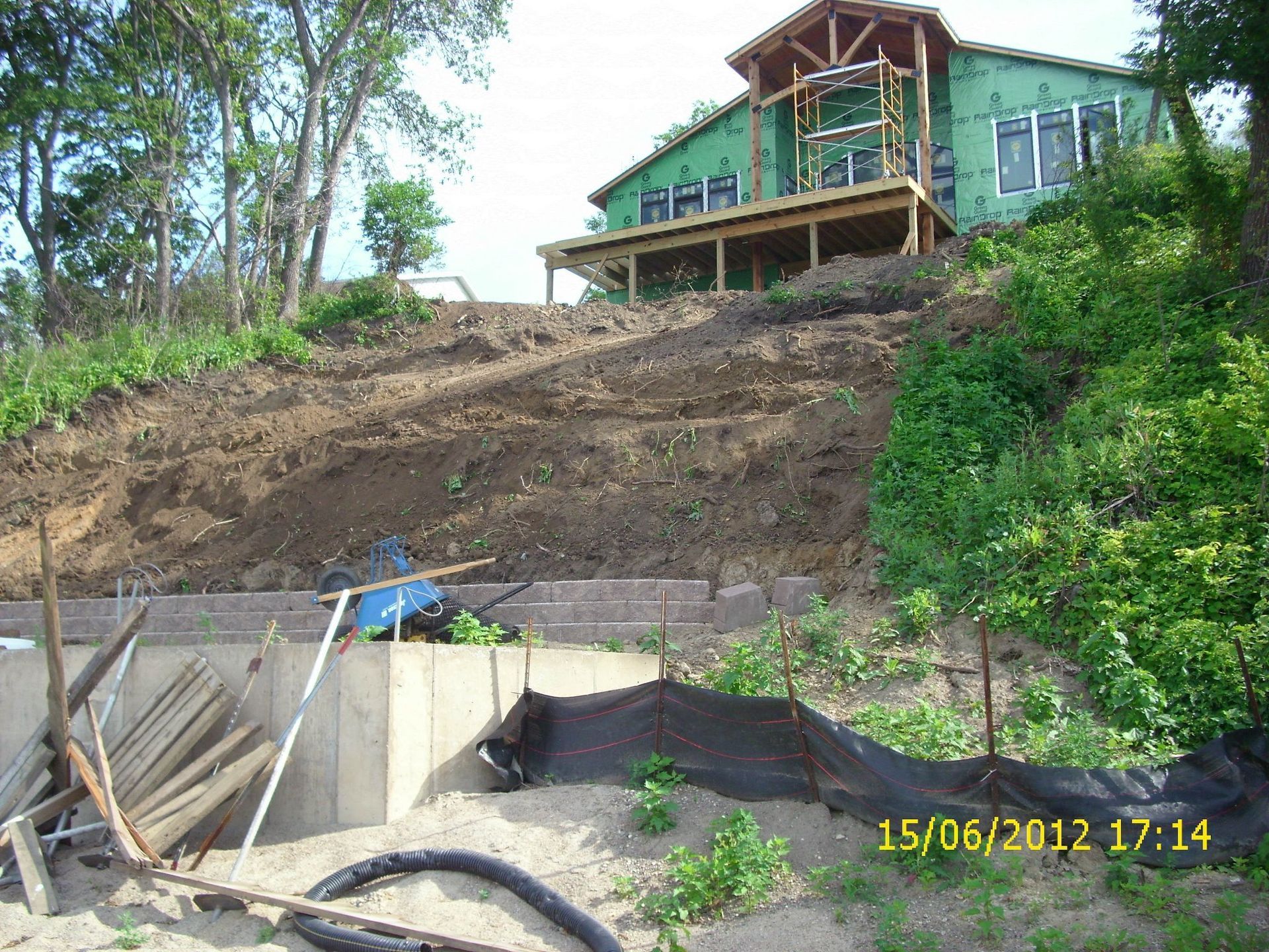 A house under construction sits atop a steep, terraced hillside with concrete retaining walls and erosion control netting.