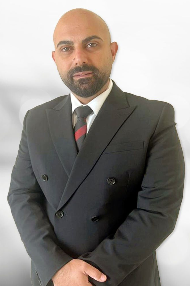 Man in dark suit, white shirt, and tie, arms crossed, standing with neutral expression.