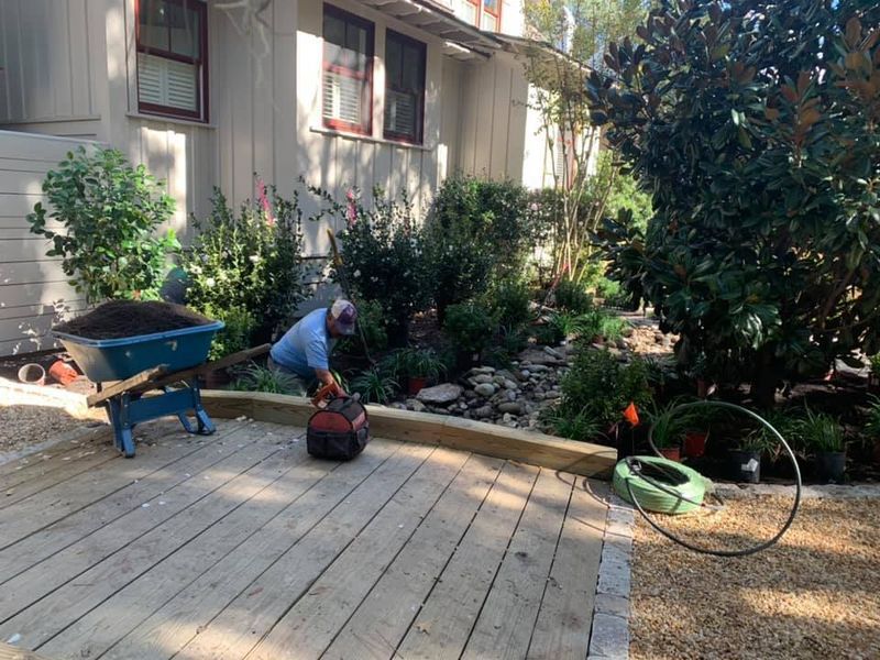 Person gardening, with wheelbarrow of soil, near a house and garden with a hose.