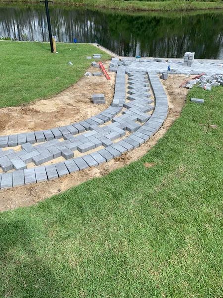 Pathway construction near a pond; gray paving stones laid in a curved design on a sandy base, surrounded by green grass.