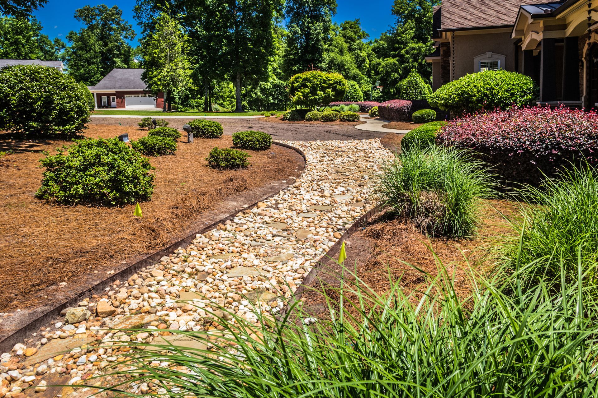 Gravel-lined stream in a landscaped yard with lush greenery, shrubs, and a house in the background on a sunny day.