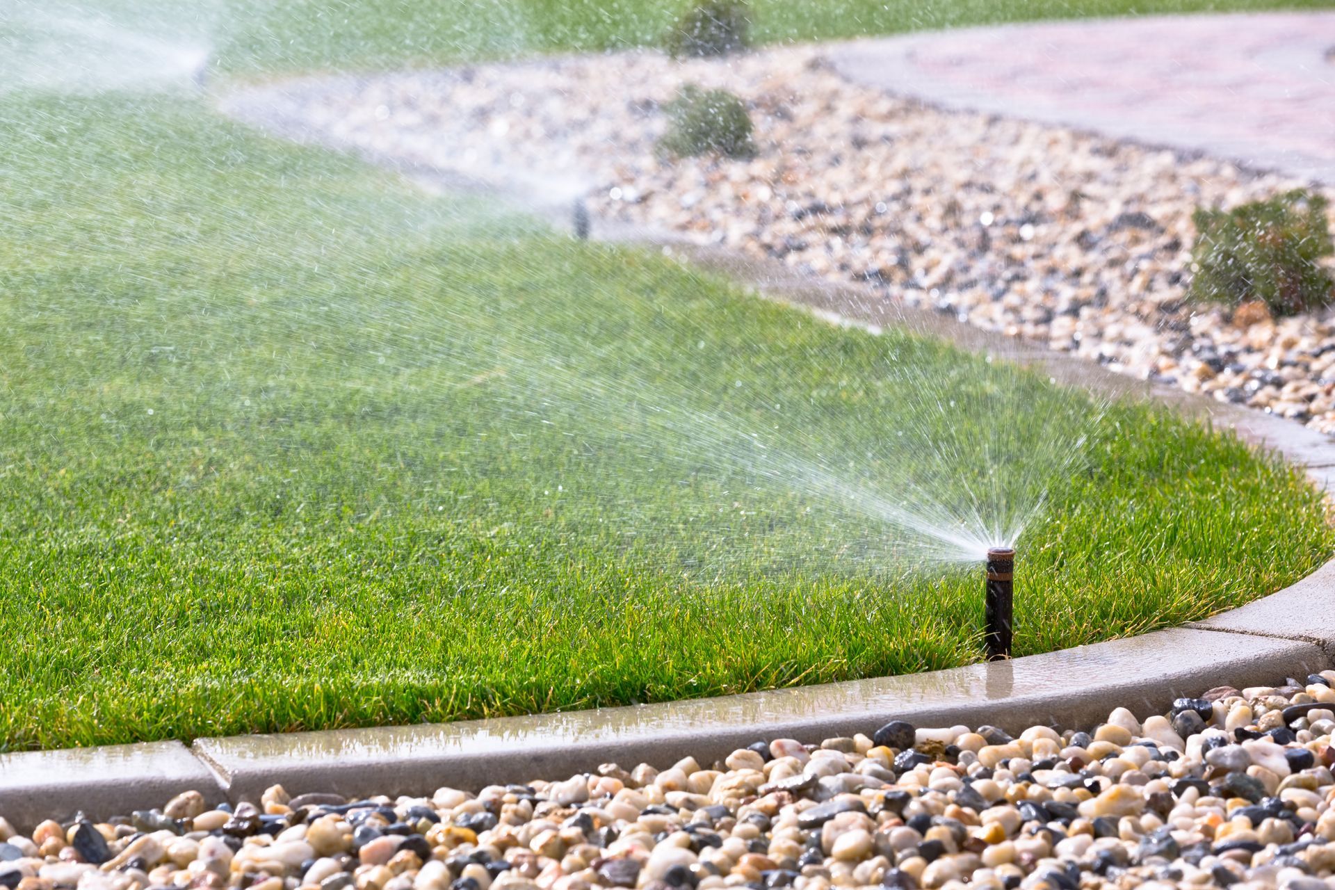 Sprinkler watering green lawn, stone border, and gravel landscaping.