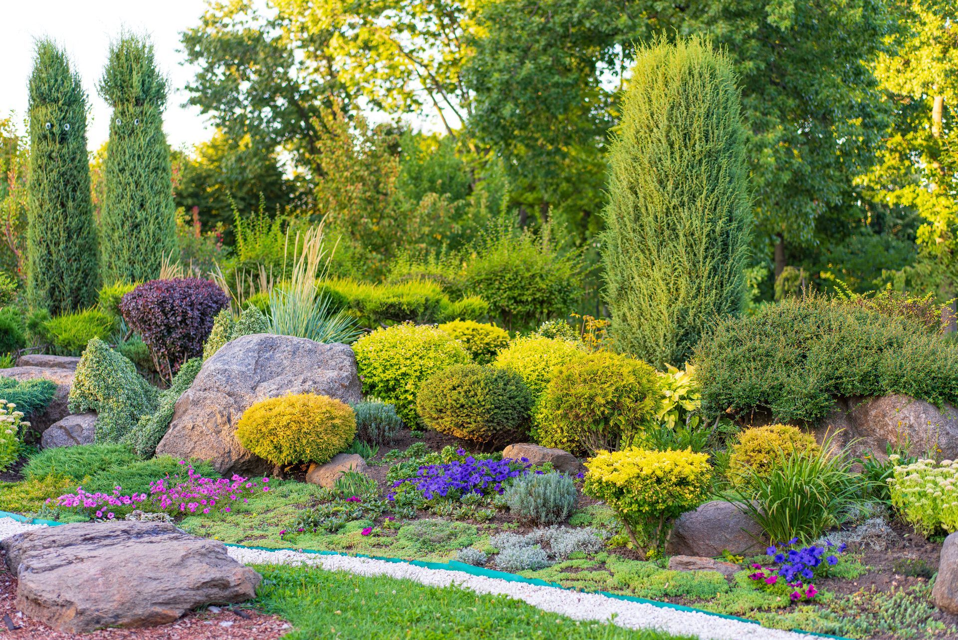 Lush garden bed with boulders, various green, yellow, and purple plants.
