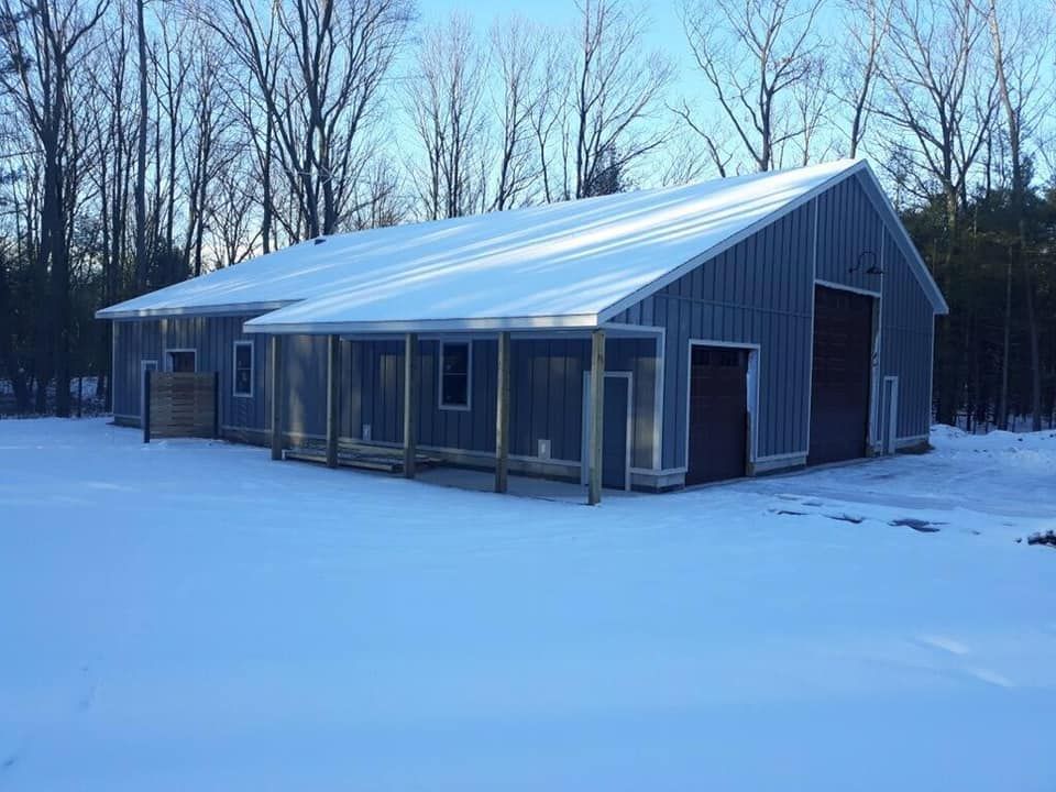 Gray barn with metal roof and porch, covered in snow, set in a winter landscape with trees.