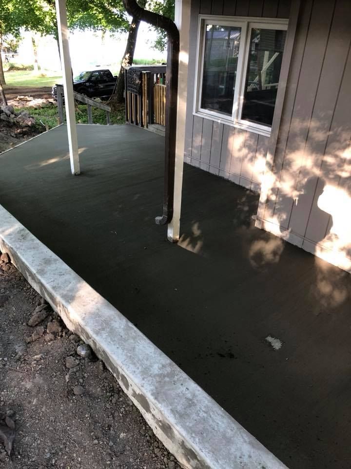 A newly poured concrete patio alongside a house with a window, downspout, and low retaining wall.