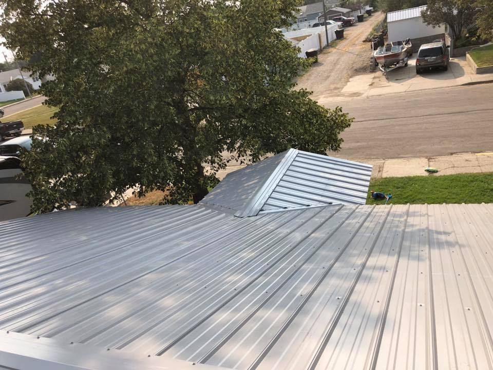 Silver metal roof with a small, gabled section, viewed from above, with a street and trees in the background.