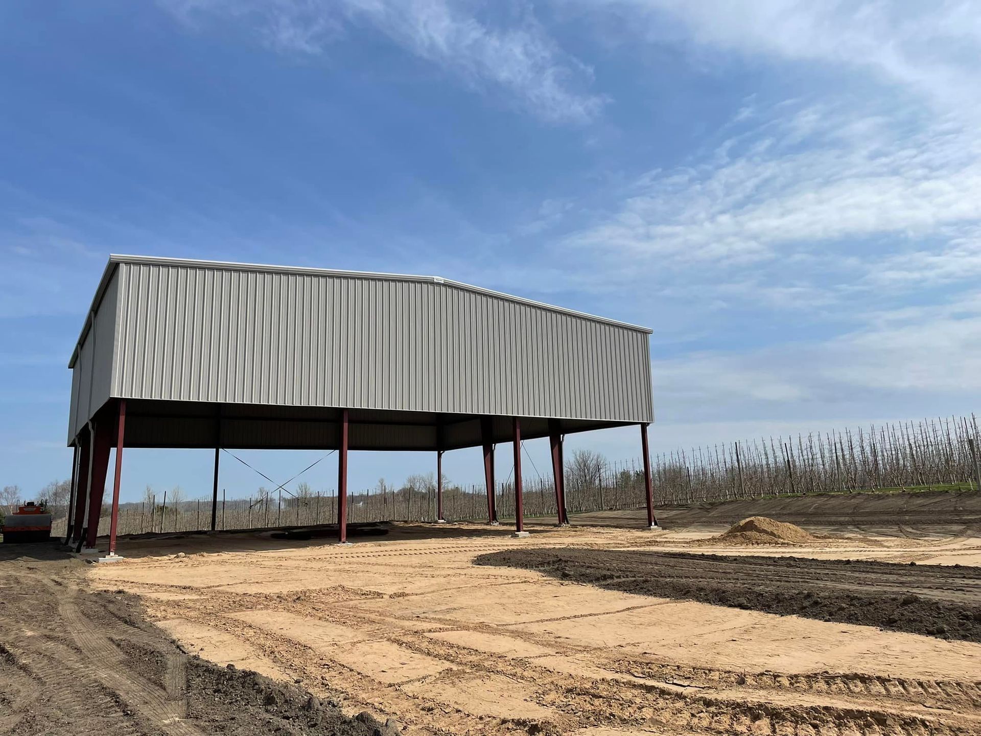 A metal shed on brown dirt, supported by red posts. Blue sky, field in background.