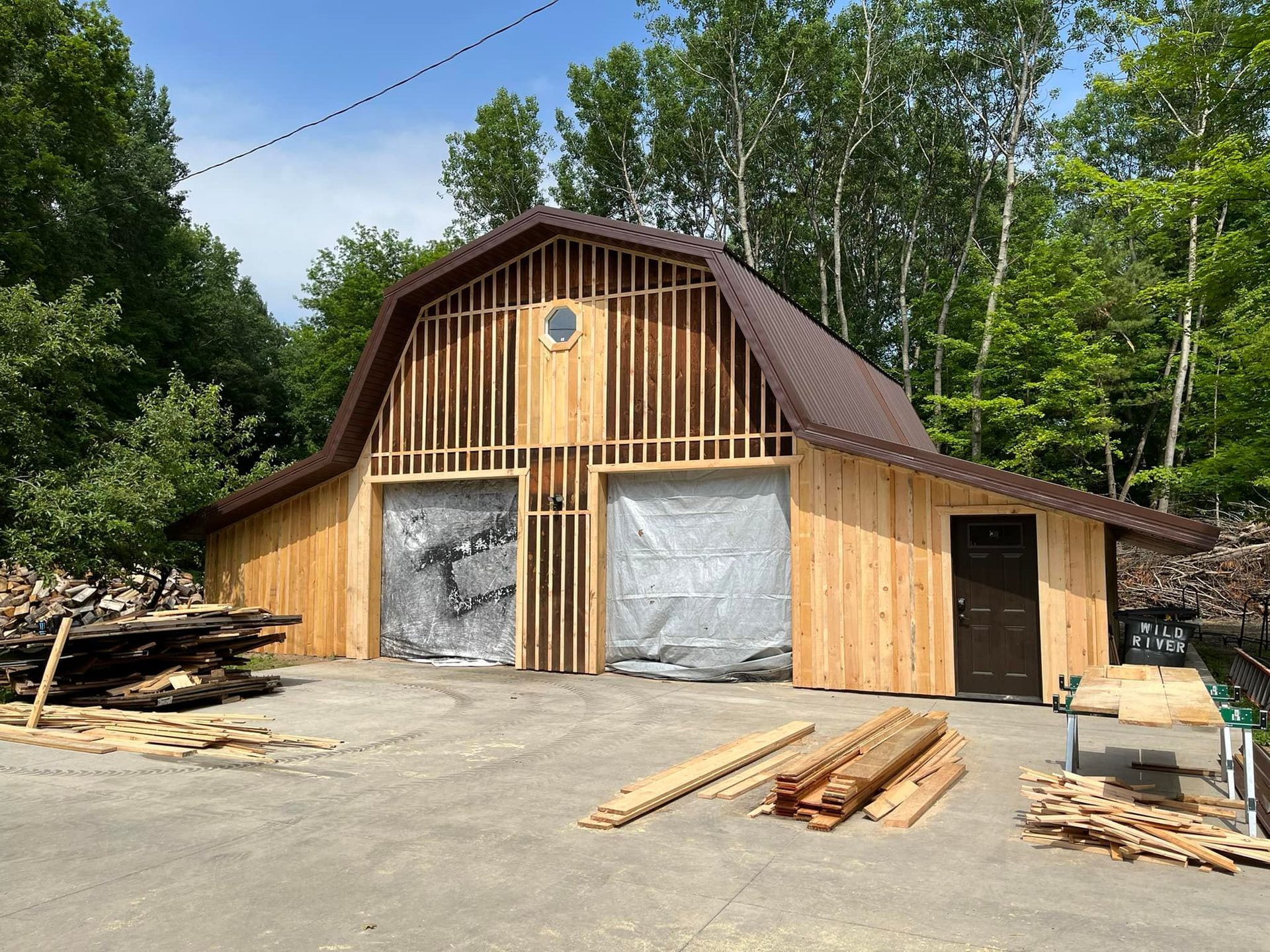 Barn under construction, wooden frame, metal roof, two garage doors, dark brown door, lumber on ground, surrounded by trees.
